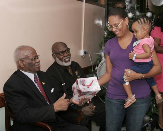 Christine Smith holds her young daughter, Zahra Smith, in her arms, as she collects a Christmas gift on behalf of her daughter from the Governor General of Barbados, His Excellency Sir Elliott Belgrave, on his annual visit to the Queen Elizabeth Hospital (QEH). Looking on is the Chairman of the QEH Board of Management, Joseph King.